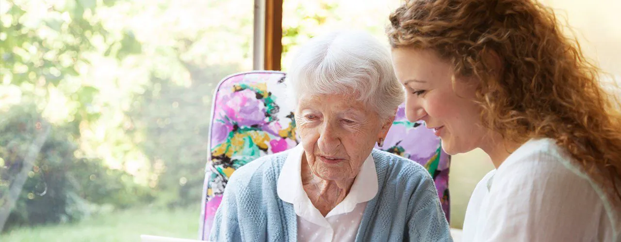 Home carer supporting an older client beside a window